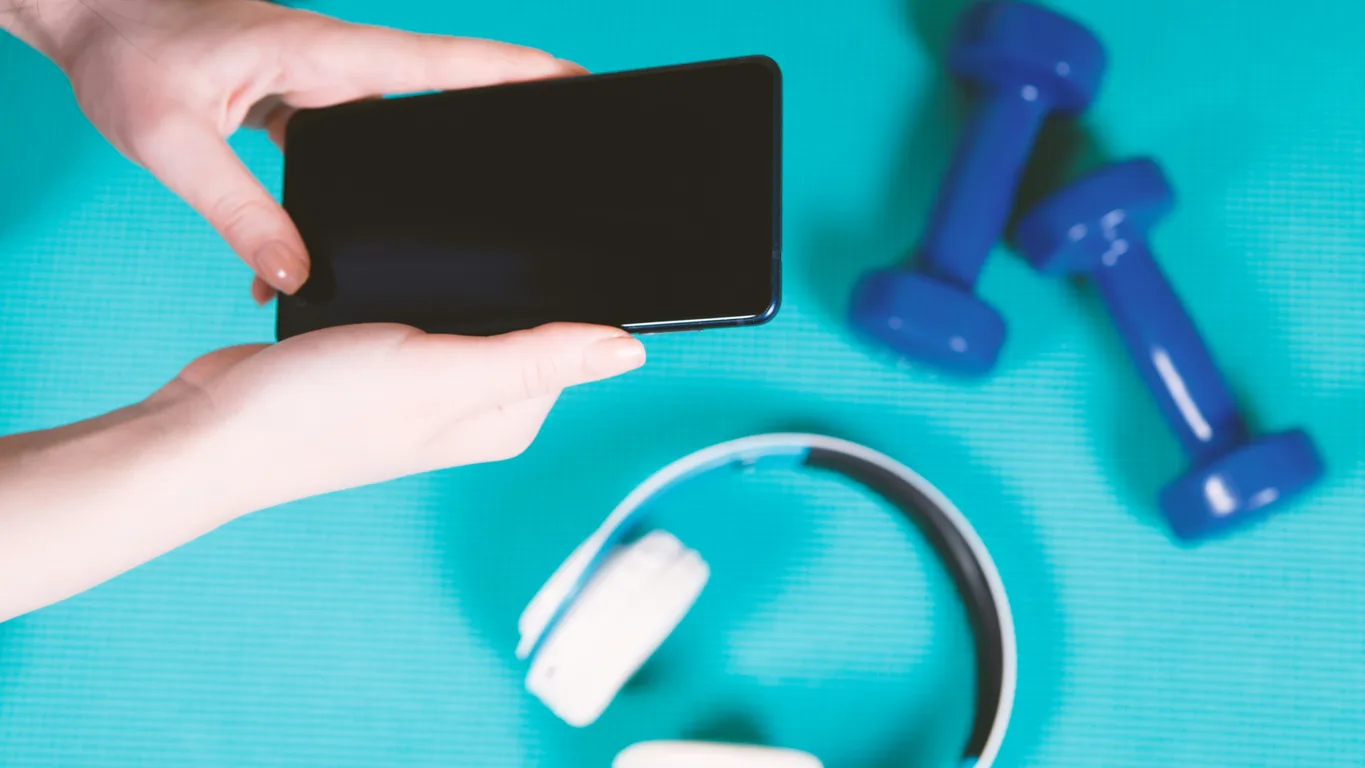 Hände halten ein Smartphone über einer türkisfarbenen Trainingsmatte, daneben Kurzhanteln und Kopfhörer