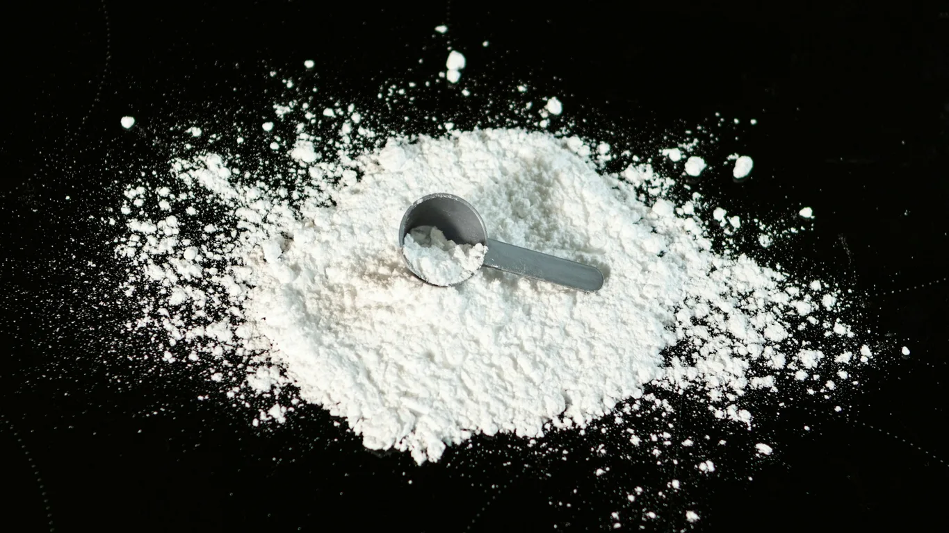 Kreatin-Pulver im Glas mit Messlöffel neben Shaker und Wasserglas in heller Küche